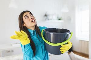 mulher chocada olha para o teto enquanto coleta água que vaza na sala de estar em casa. mulher preocupada segurando balde enquanto gotas de água vazam do teto na sala de estar foto