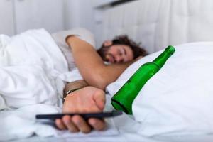 mensagens de texto enquanto conceito bêbado. jovem deitado na cama mortalmente bêbado segurando uma garrafa vazia de bebida. embriagado com álcool. alcoolismo embriaguez habitual conceito de hábito pernicioso foto