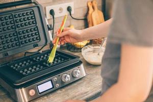 jovem mulher preparando waffles belgas na cozinha, untando o waffle iron com óleo de coco. processo de cozimento foto