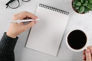 as mãos da mulher escrevendo para fazer a lista ou planos no caderno e segurando a xícara com café na mesa do escritório. vista superior, copie o espaço foto