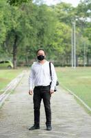 homem asiático freelancer de cabelos longos com máscara facial preta está de pé e postando no caminho a pé no campo ao ar livre do jardim com sua câmera ao lado dele. foto