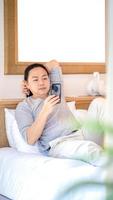 relaxante e feliz homem asiático de cabelo comprido deitado na cama e jogando móvel pela manhã. foto