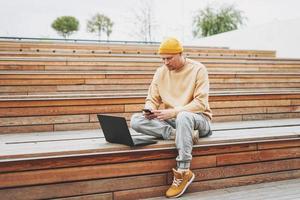 hipster de homem elegante em freelancer de chapéu amarelo trabalhando no laptop no parque da cidade de rua foto