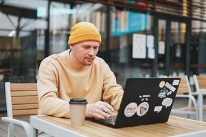 hipster de homem elegante em freelancer de chapéu amarelo trabalhando no laptop no café da rua foto