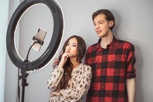 jovem casal de blogueiros de beleza, cabeleireiro, cabeleireiro e modelo com fotos de estilo de cabelo no celular com lâmpada de anel no fundo da parede cinza