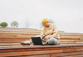 hipster de homem estiloso em freelancer de chapéu amarelo trabalhando em laptop no parque da cidade de rua