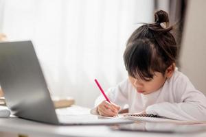 colegial asiática fazendo sua lição de casa com o laptop em casa. as crianças usam aparelhos para estudar. educação e ensino a distância para crianças. educação domiciliar durante a quarentena. ficar em casa foto