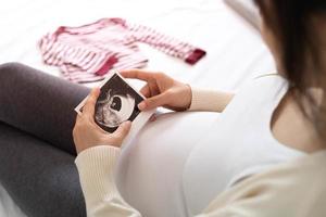 uma mulher grávida está olhando para uma foto de ultrassom do feto. a mãe toca suavemente o bebê no estômago. as mulheres estão grávidas por 30 semanas. primeiro amor na barriga e última gravidez