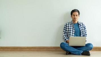 homem asiático vestindo uma camisa xadrez azul trabalhando em um laptop, relaxando no chão da sala dentro de casa. homem asiático, trabalho em casa conceito foto