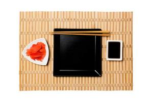 prato quadrado preto vazio com pauzinhos para sushi, gengibre e molho de soja no fundo amarelo da esteira de bambu. vista superior com espaço de cópia para você projetar foto