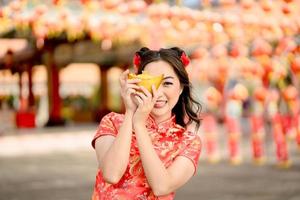 jovem mulher asiática usando vestido cheongsam qipao tradicional segurando dinheiro de ouro antigo no templo budista chinês. comemore o ano novo lunar chinês, feriado da temporada festiva. sorriso de emoção foto