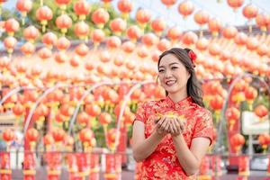 jovem mulher asiática vestindo traje tradicional cheongsam qipao segurando dinheiro de ouro antigo no templo budista chinês. conceito de ano novo chinês. sorriso de emoção foto
