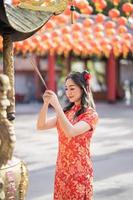 imagem vertical. linda mulher asiática vestindo vestido cheongsam qipao tradicional rezando com paus de incenso durante o ano novo chinês ou lunar, ano novo chinês foto