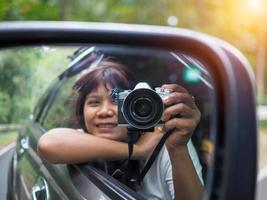uma mulher segura uma câmera digital e tira uma foto de si mesma sorrindo refletida no retrovisor do carro.