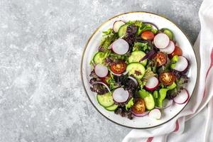 salada de legumes saudável de tomate cereja, fatias de pepino, folhas de alface verde e roxa, cebola e azeite no prato na mesa de concreto vista plana dieta leiga, menu mediterrâneo comida vegana foto