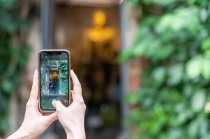 mão feminina usa smartphone para tirar foto com a estátua de Buda de ouro em uma igreja coberta com figueira-de-bengala de 100 anos em wat bangkung.