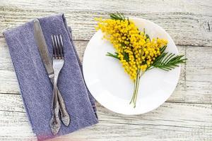 prato de configuração de mesa festiva com garfo de guardanapo e faca e decoração de flores de mimosa em fundo branco de madeira foto
