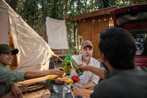 amigos de grupos de turistas acampando e fazendo um churrasco e bebendo cerveja-álcool e quebrando uma garrafa de cerveja com a festa junto com diversão e felicidade no verão foto
