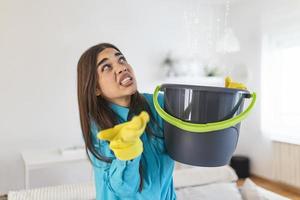 mulher chocada olha para o teto enquanto coleta água que vaza na sala de estar em casa. mulher preocupada segurando balde enquanto gotas de água vazam do teto na sala de estar foto