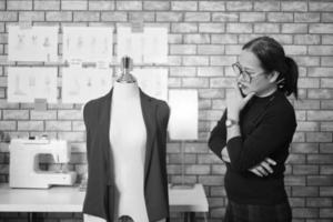 retrato em preto e branco, designer de moda feminina madura asiática fica perto de fantoche de vestir, pensando e contemplando ideias de design, trabalhos de coleção de roupas, alfaiate boutique profissional. foto