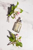 um frasco com um cosmético ou soro para o cuidado da pele do rosto em um fundo branco de mármore e galhos de uma macieira em flor. vista do topo.