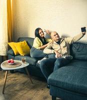 casal usa dispositivo de smartphone, sentado no apartamento aconchegante do sofá. tire selfie juntos como namorado e namorada. fazer bate-papo por vídeo na internet, usar mídias sociais, transmitir vídeo ao vivo