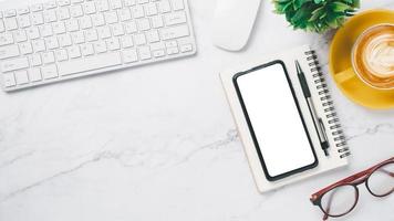 espaço de trabalho de mesa de escritório branco com telefone inteligente de tela em branco, teclado de computador, mouse, óculos, caneta e xícara de café, vista superior plana com espaço de cópia. foto