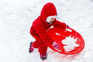menina muito sorridente em seu traje de esqui deslizando por uma pequena colina coberta de neve com seu trenó foto