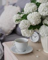 uma xícara de café, um despertador e um vaso de flores brancas na mesa de cabeceira. hora do chá, hora do café foto