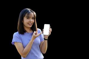 mulher asiática sorridente jogando e atualizando novos aplicativos. sua mão apontada para o telefone com área de trabalho de tela em branco conversando sobre fundo preto. foco suave e seletivo. foto