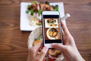 a mão de uma mulher usando um celular para tirar fotos de comida na mesa para revisar e enviar para as redes sociais