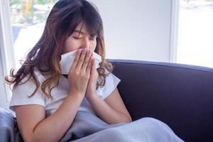 mulher de cabelos compridos sentada no sofá está sofrendo de gripe, tosse e espirros. sentado em um cobertor por causa da febre alta e cobre o nariz com lenço de papel porque espirra o tempo todo. foto