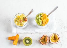 dois copos com iogurte e purê de frutas e com colheres metálicas e meias frutas em fundo branco foto