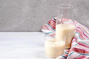 vidro de close-up e garrafa com leite de aveia e toalha de cozinha listrada na mesa de madeira branca no fundo da parede de concreto. foto