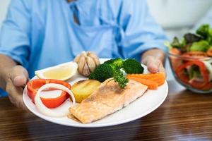 paciente senhora idosa asiática ou idosa comendo o pequeno-almoço de filé de salmão com alimentos vegetais saudáveis enquanto está sentado e com fome na cama no hospital. foto