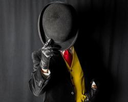 retrato de homem de terno escuro e colete amarelo e gravata vermelha segurando chapéu-coco sobre o rosto foto