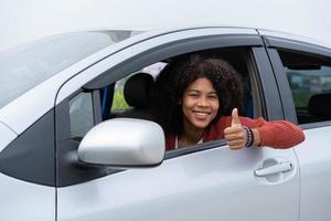 jovem meio asiático meio africano mostra o polegar para cima dentro do novo carro moderno. jovem emoção primeira viagem depois de passar no teste de carteira de motorista. se sente bem depois de comprar um carro novo. foto