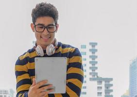 o homem de óculos de negócios criativos jovens usa fones de ouvido e se sente feliz enquanto olha para o tablet para trabalhar com um rosto sorridente na frente do fundo da janela do escritório. fotografia de luz de fundo.