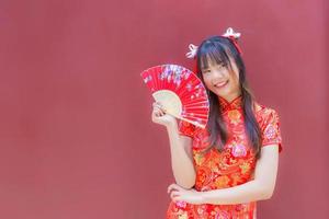 bela jovem asiática no vestido cheongsam vermelho no tema do ano novo chinês fica sorrindo alegremente olhando para a câmera segura um leque sobre fundo vermelho. foto