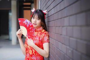 linda mulher asiática de vestido vermelho fica sorrindo alegremente olhando para a câmera segura um leque entre o antigo centro da cidade no tema do ano novo chinês. foto