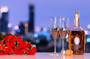 seletivo no primeiro copo de vinho espumante rosé com garrafa turva e rosas vermelhas na luz colorida bokeh do fundo da cidade para o conceito de jantar dos namorados. foto
