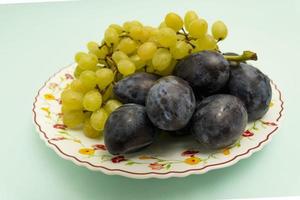 uvas brancas orgânicas maduras com ameixas orgânicas azuis redondas em um prato sobre fundo turquesa, frutas, alimentos saudáveis foto