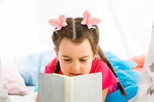 curtindo uma ótima história. menina bonitinha lendo livro enquanto está sentado no chão na barraca foto