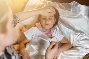 menina doente na cama. mãe com termômetro está sentada perto da cama. foto