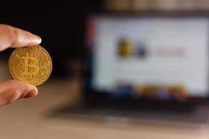 moeda de ouro bitcoin na mão do homem no fundo do laptop em uma mesa branca foto