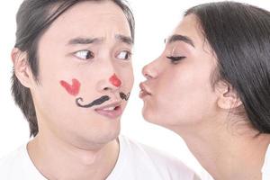 casal misto se divertindo. modelo feminino árabe dando beijo ao modelo masculino asiático que afogou bigodes e coração no rosto. isolado no fundo branco. foto