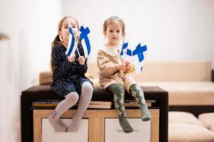 duas irmãs estão sentadas em um sofá em casa com bandeiras finlandesas nas mãos. Finlândia crianças meninas com bandeira. foto