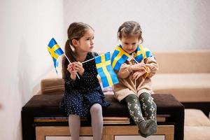 duas irmãs estão sentadas em um sofá em casa com bandeiras suecas nas mãos. Suécia crianças meninas com bandeira. foto