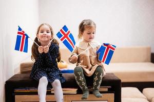 duas irmãs estão sentadas em um sofá em casa com bandeiras islandesas nas mãos. Islândia crianças meninas com bandeira. foto