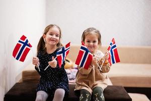 duas irmãs estão sentadas em um sofá em casa com bandeiras norueguesas nas mãos. noruega crianças meninas com bandeira. foto